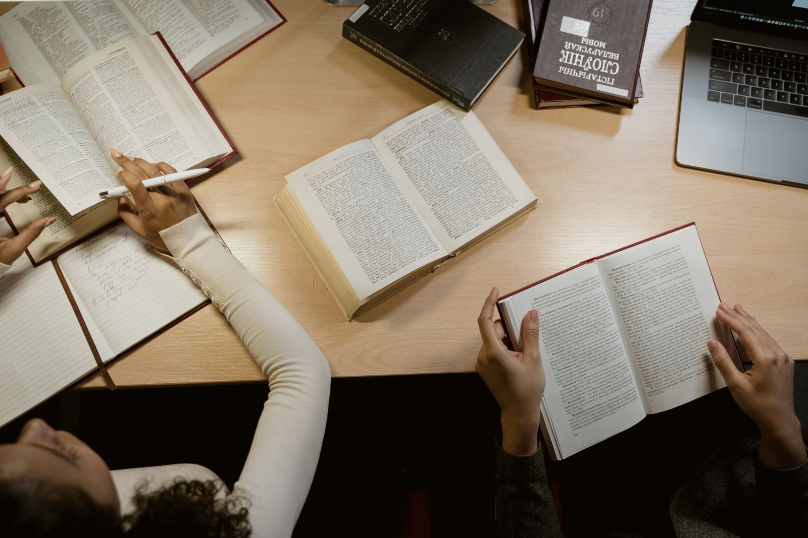 木のテーブルの上に開いた本とノートパソコンを囲んで勉強する人たち